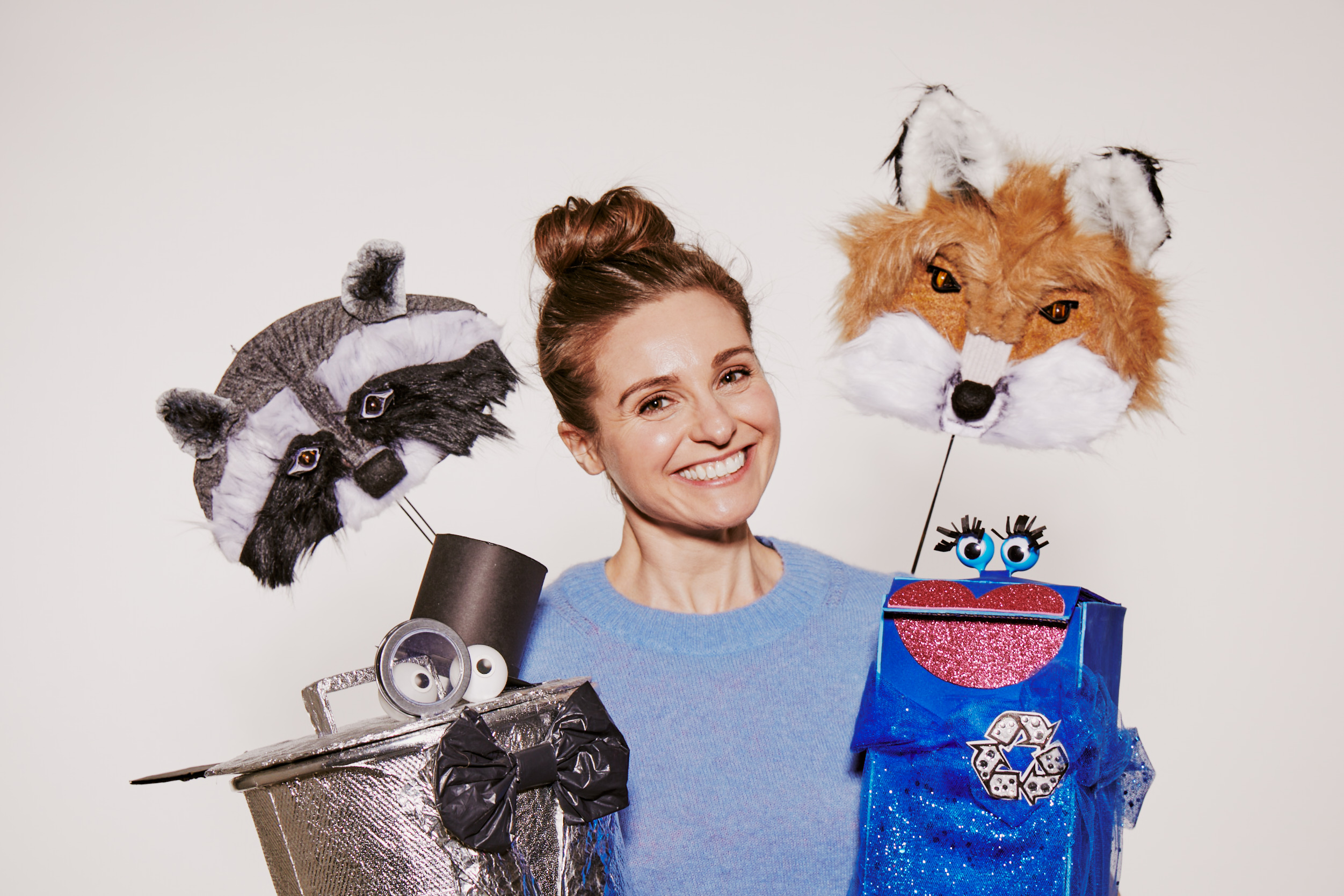 Betsy Rosen wearing a light blue sweater is holding two puppet heads on sticks. One is a raccoon puppet with a gray and black mask, and the other is a fox puppet with orange and white fur. 
