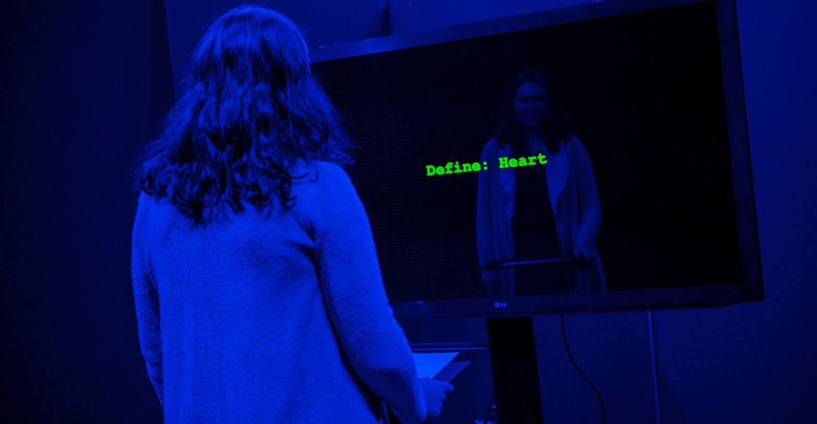 A woman in a dark exhibit reads a prompt on a screen that says define heart.