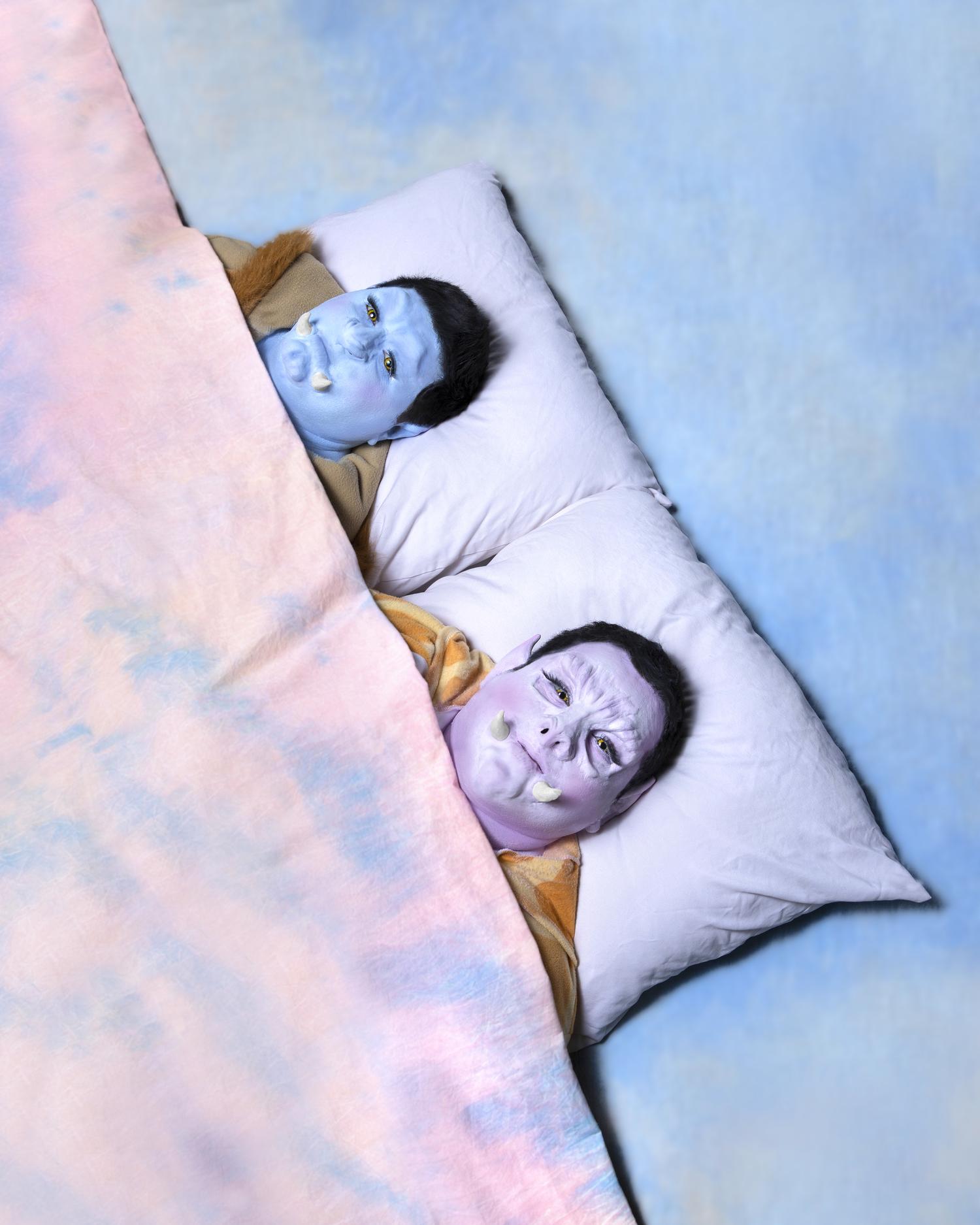 Two men wearing animal-like masks with tusks lie in a blue and pink pastel-colored bed.