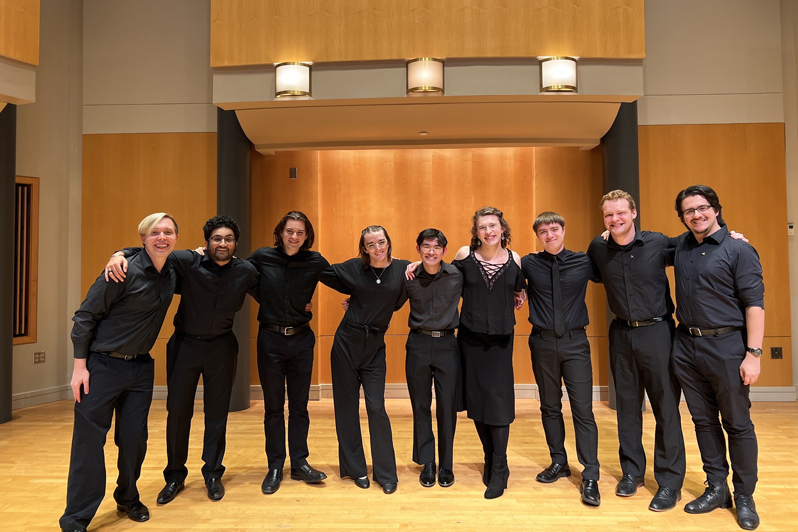 A capella members in all black concert attire pose for a photo in a concert hall.