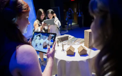 Students gather around a table, looking at a tablet that scans a small model with houses, trees, and buildings.