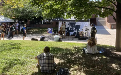 Students sit in the grass watching a live performance at the Arts for All tent at the UMD Farmers Market.