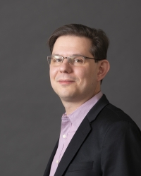 A man looks at the camera almost in profile. He wears glasses, a light purple shirt and a black suit jacket.