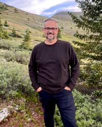 A man looks at the camera and smiles. He is outside surrounded by a hill and trees.