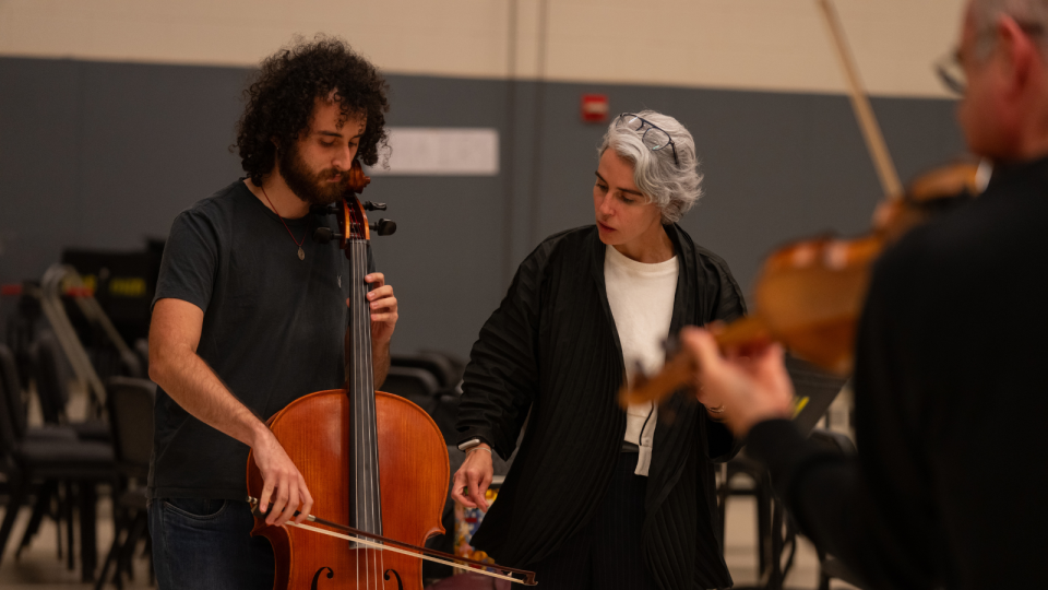 Clarice Assad works with John Pablo Rojas. He is holding his cello, and both are looking down at the instrument.