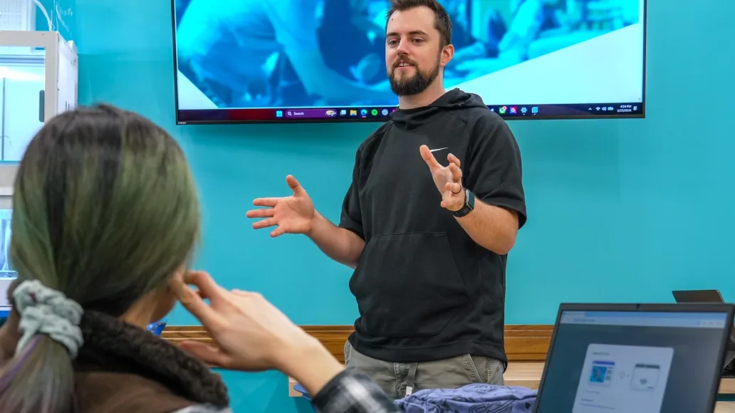 A man is speaking in front of a screen to a woman who is looking on.
