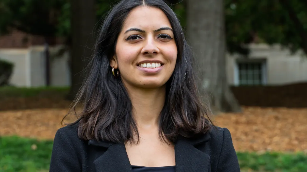 A woman looks at the camera smiling. There is a tree and grass behind her.