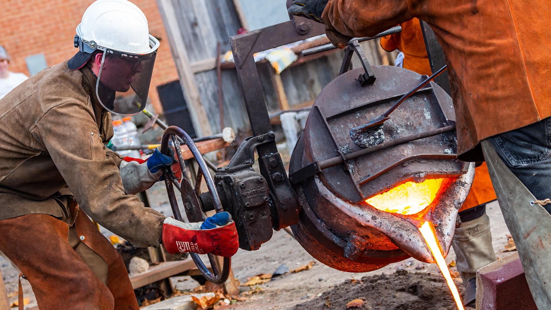 Student pours molten metal from a bucket-like furnace.