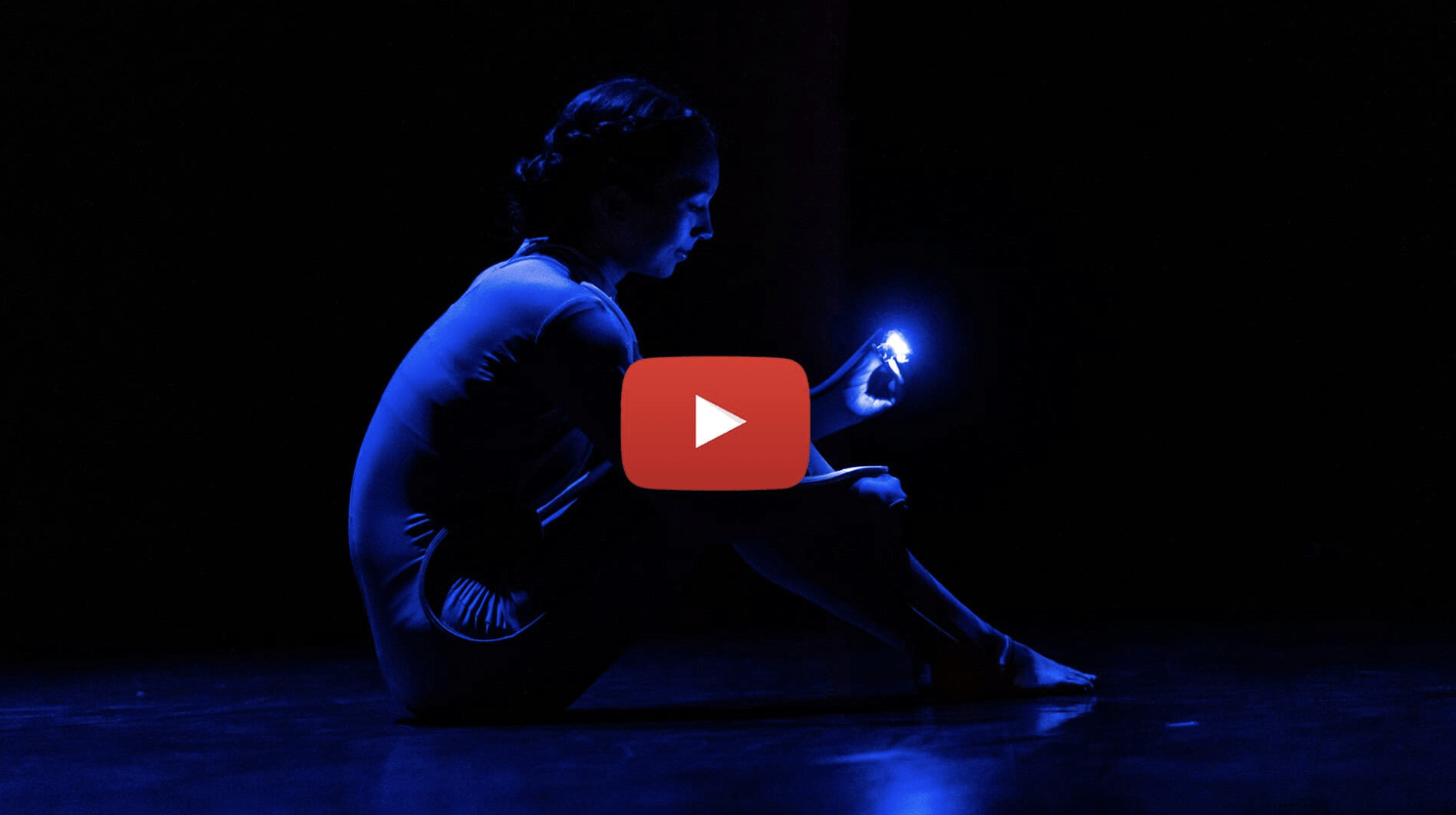 A dancer sits on a dark stage with an electric sensor on her arm.